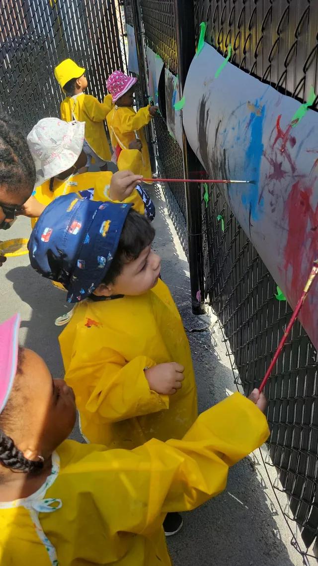 Des enfants en tabliers jaunes peignent sur une grande feuille accrochée à la clôture